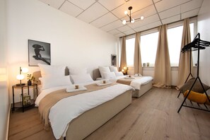 A hotel room with two beds, a bedside table with a lamp, a wall-mounted artwork, and a window with curtains.