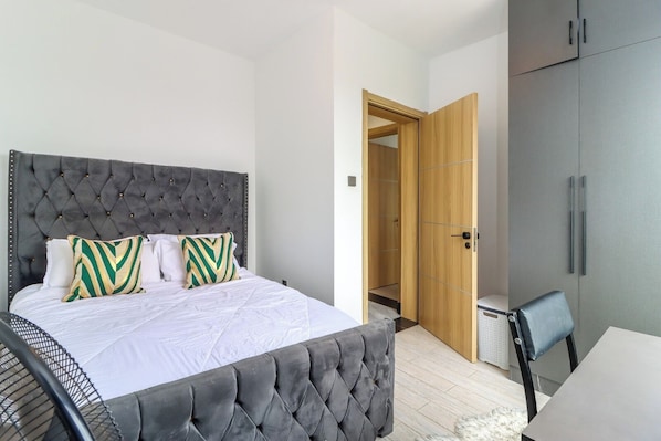 A bedroom with a tufted headboard, a bed with white bedding, two patterned pillows, a grey upholstered bench, a wooden wardrobe, and a small table with a chair.