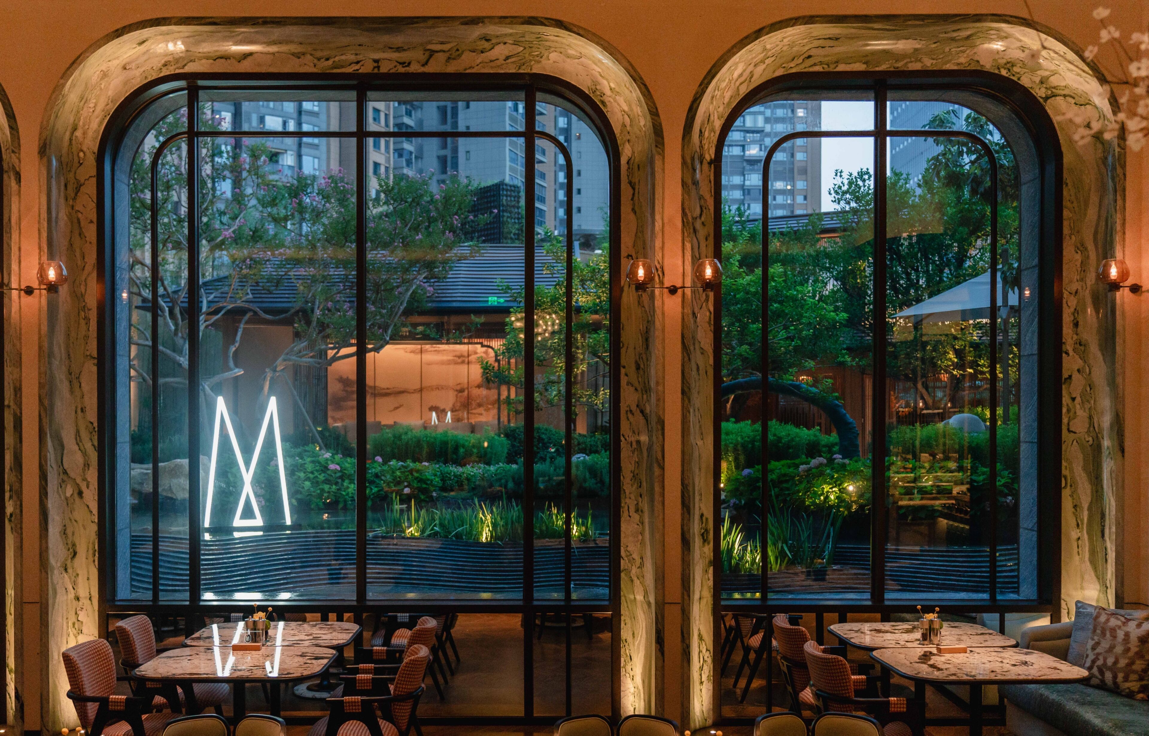 Un restaurante con amplias ventanas arqueadas, mesas dentro y un jardín visible al aire libre.