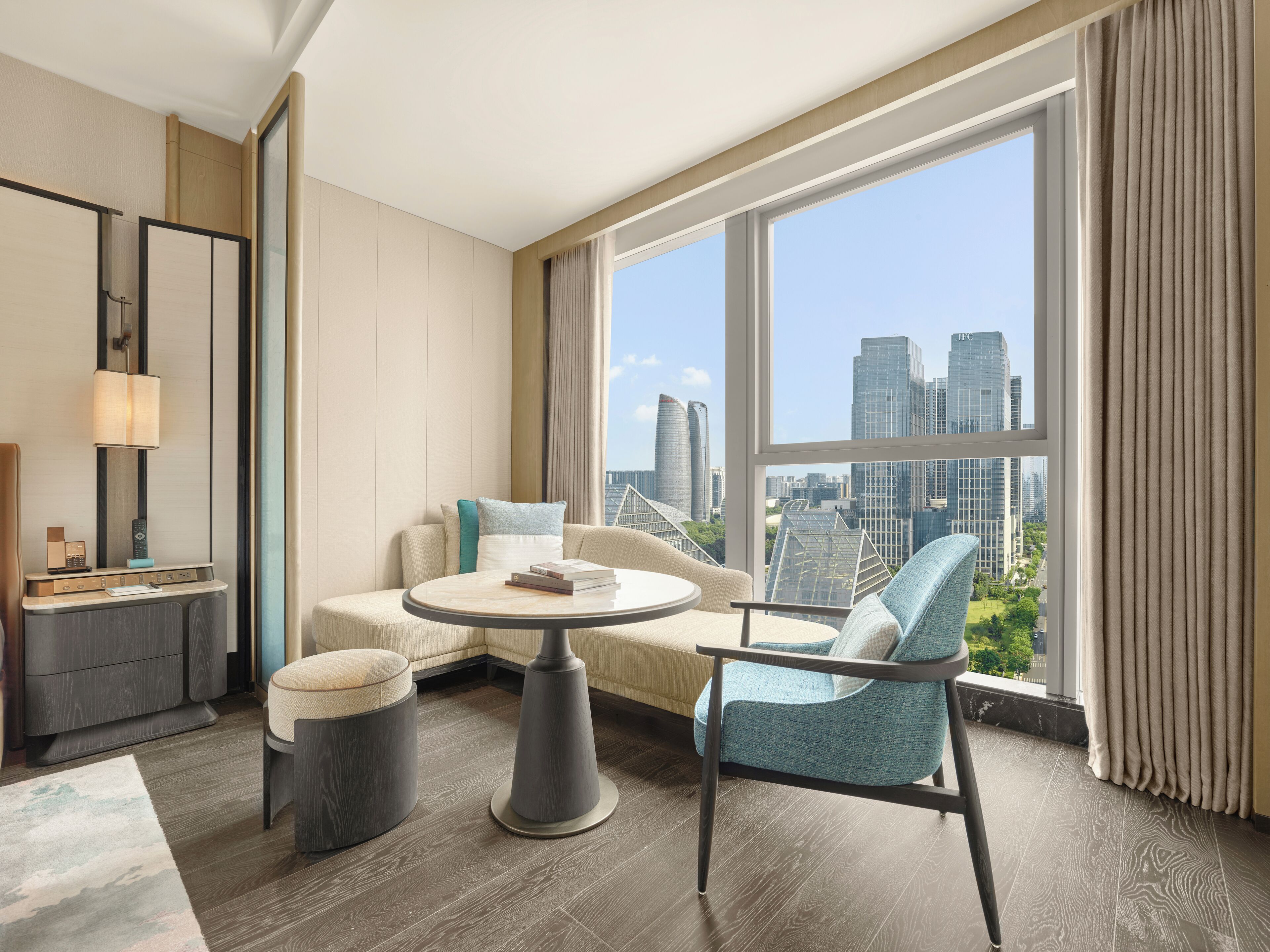 A modern hotel room with a round table, a sofa, a chair, and a large window offering a city view.