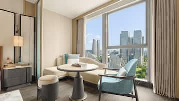 A modern hotel room with a round table, a sofa, a chair, and a large window offering a city view.