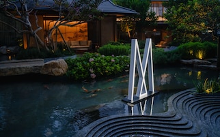 A koi pond with a modern sculpture, surrounded by well-maintained greenery and a traditional building in the background.