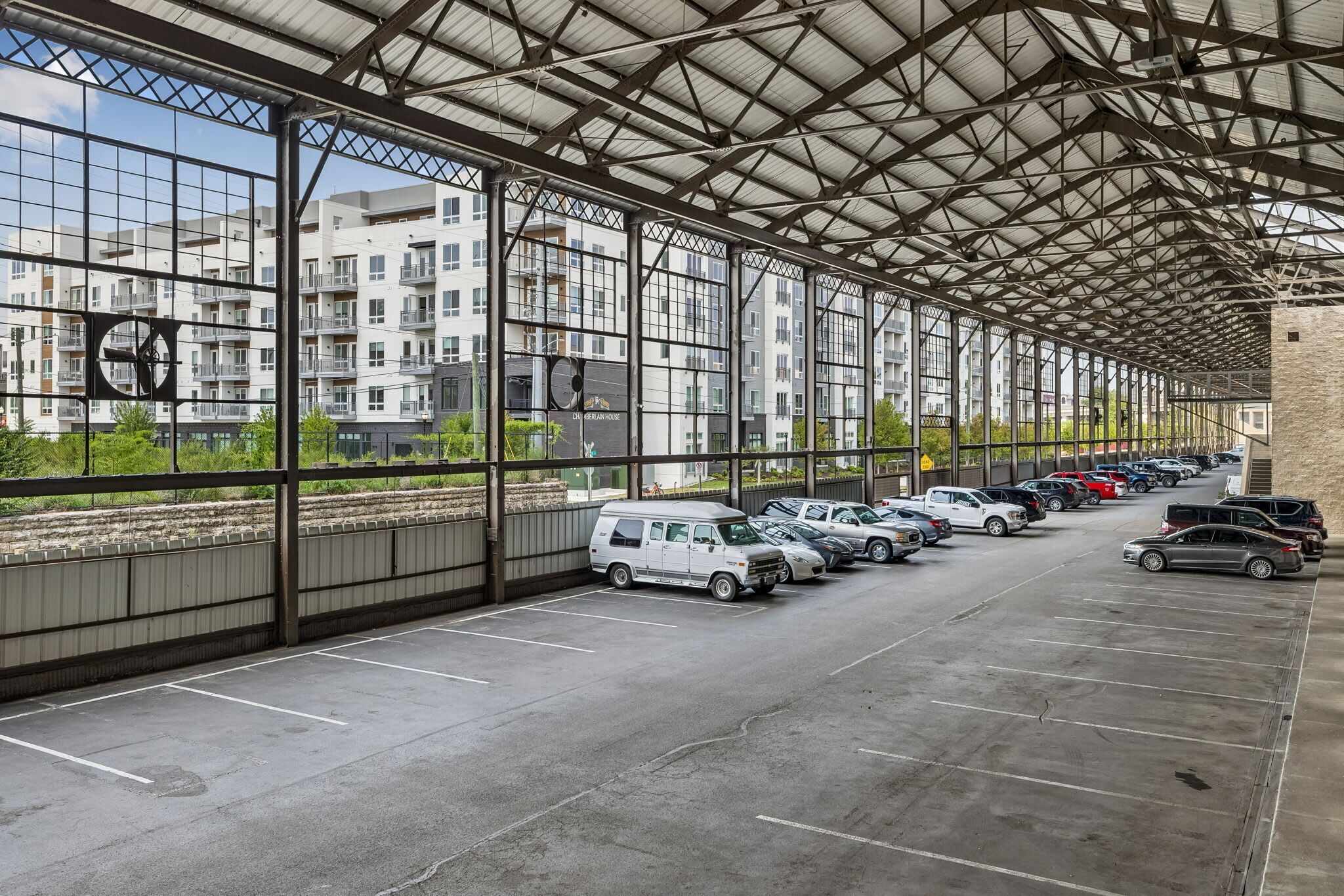 A covered parking area with multiple rows of parked cars and a view of residential buildings in the background.