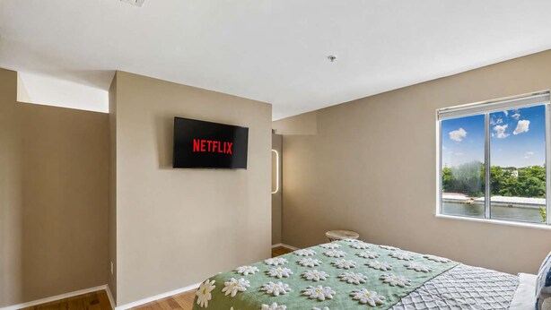 Un dormitori amb un llit, una televisió penjada a la pared que mostra 'NETFLIX', una finestra amb vista a vegetació i una taula petita.