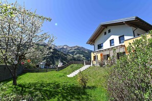 Ein modernes Haus mit einer Treppe, die zu einer Holzterrasse führt, umgeben von üppiger Vegetation und einer Bergkulisse.