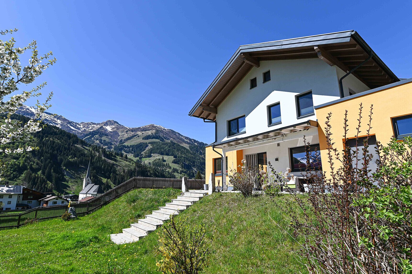 Une maison moderne à deux étages, dotée d’un toit en pente, d’un escalier en bois menant à l’entrée et offrant une vue sur une chaîne de montagnes en arrière-plan.