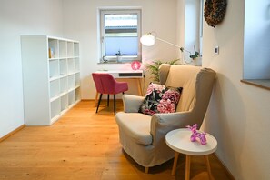 A living room with a beige armchair, a pink chair, a white side table, and a small white table with a pink figurine.