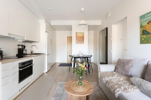 A modern kitchen with white cabinets, a dining area with a black table and chairs, and a living room with a grey sofa.