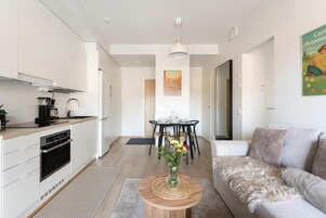 Una cocina moderna con gabinetes blancos, un comedor con mesa y sillas negras, y una sala de estar con sofá gris.