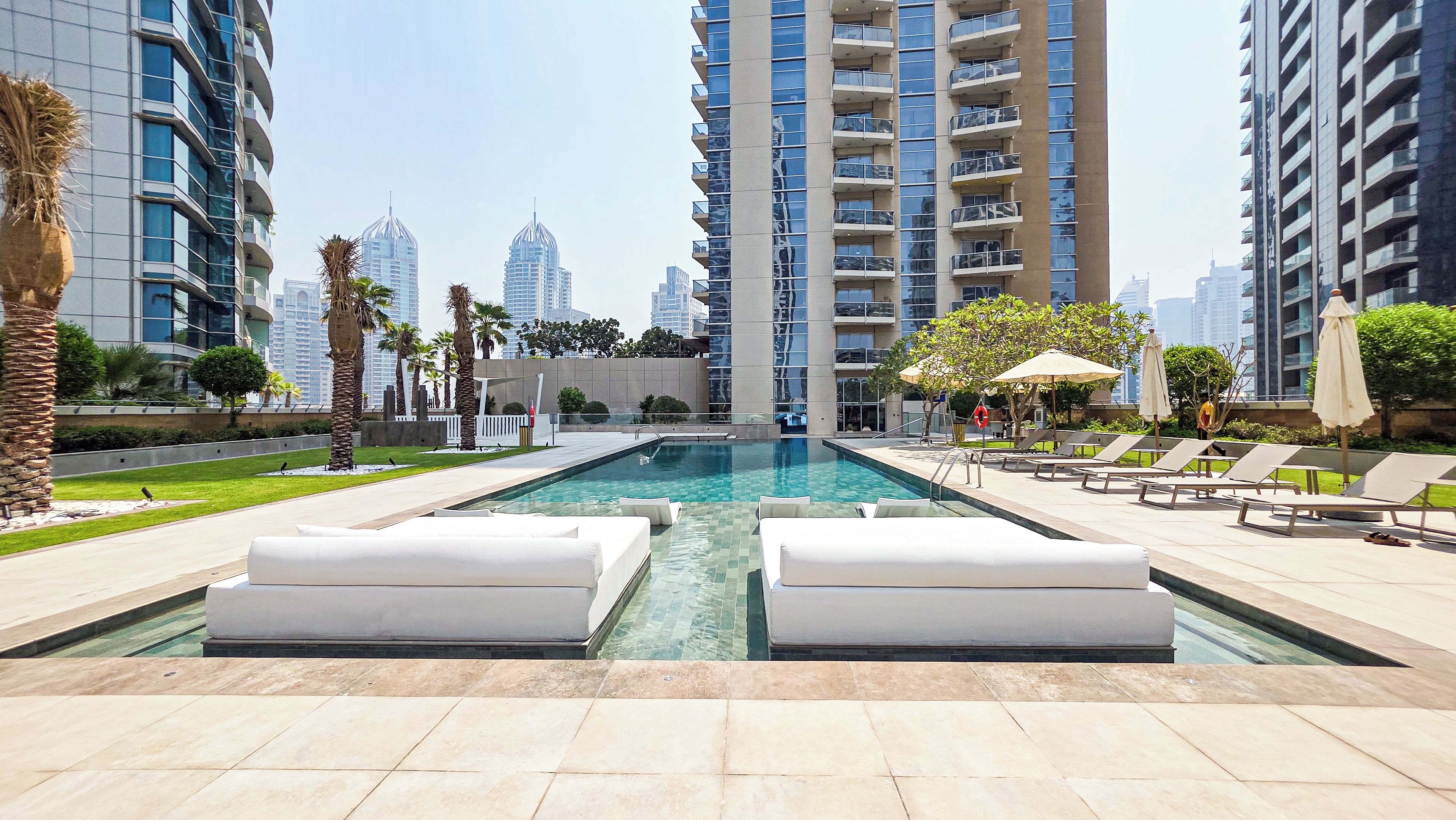 A swimming pool with lounge chairs and umbrellas, surrounded by tall buildings and palm trees.