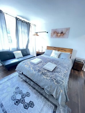 Un dormitorio moderno con cama, sofá, cabecera de madera, mesita de noche y un gran ventanal con cortinas.