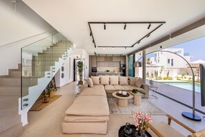 Ein zeitgemäßer Wohnraum mit Treppe, einer Sofagarnitur, einer Küchenzeile und Blick auf einen Swimmingpool.