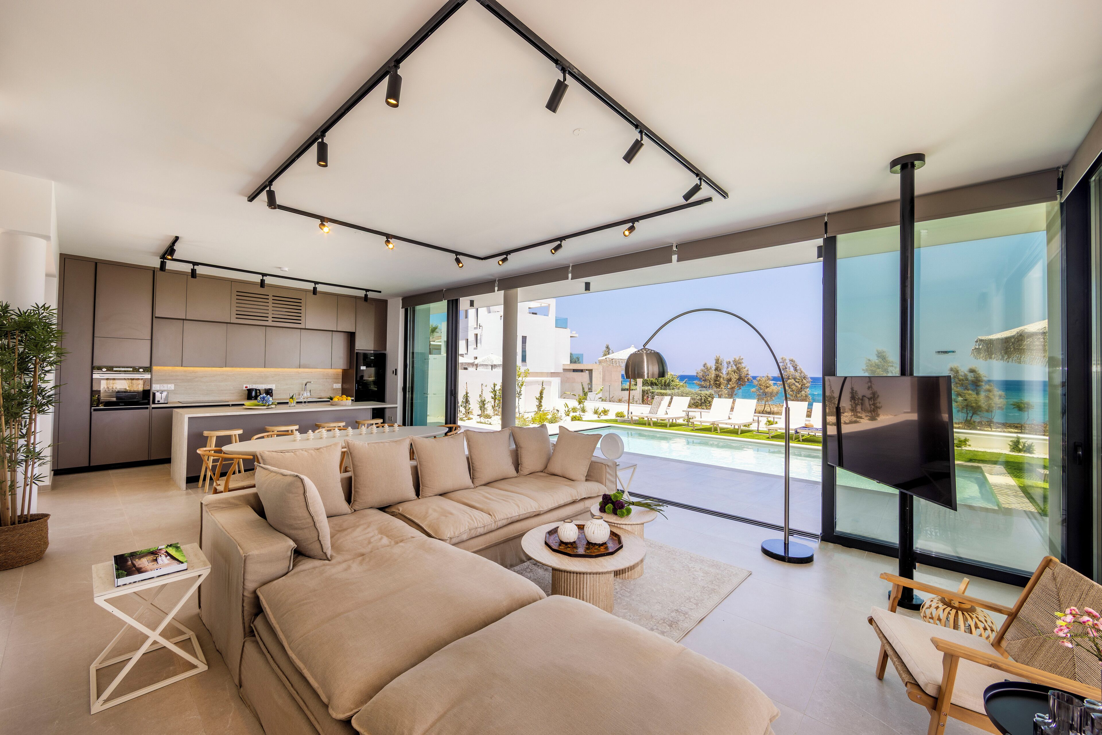 Ein modernes Wohnzimmer mit einem großen, beigefarbenen Ecksofa, einem Couchtisch und einem Fernseher. Große Fenster bieten Blick auf einen Pool und einen Strand.