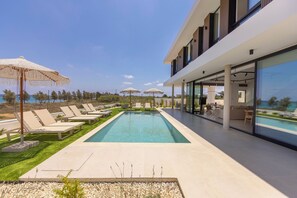 Un área de piscina moderna con sillas de descanso y sombrillas, vista despejada al mar y un edificio contemporáneo con amplias ventanas.