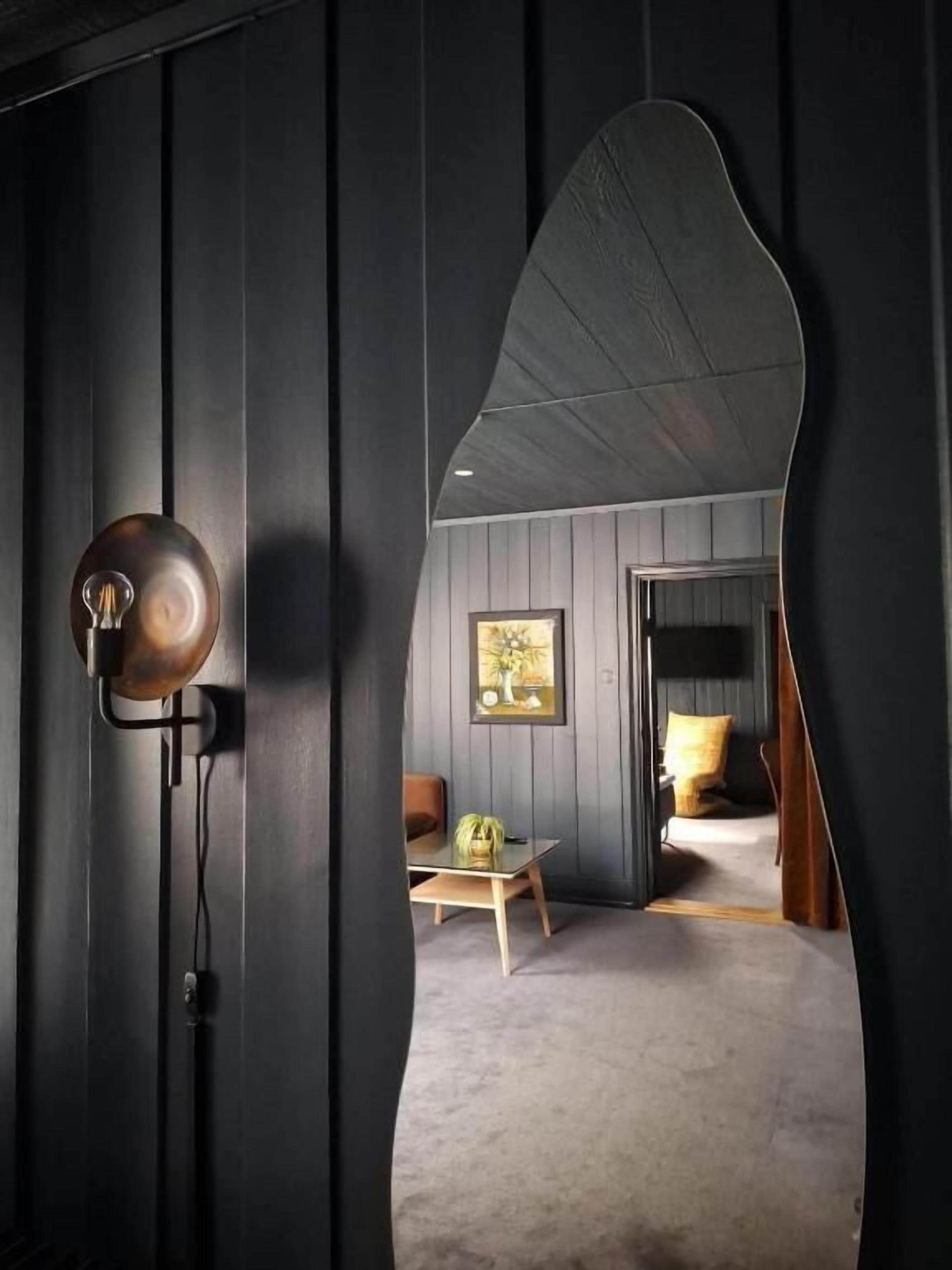 Une pièce avec un miroir incurvé, une lampe, un tableau encadré, un lit et une petite table sur laquelle se trouve un vase.