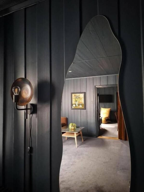 A room with a curved mirror, a lamp, a framed picture, a bed, and a small table with a vase.