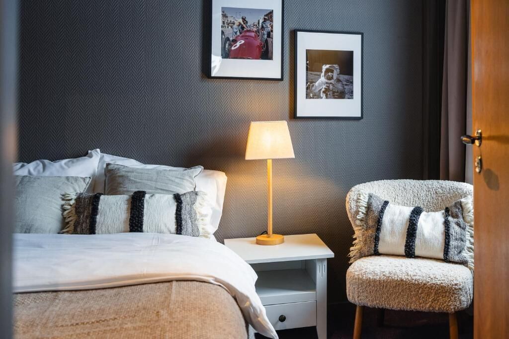 Ein Hotelzimmer mit Bett, Nachttisch mit Lampe, Stuhl und zwei gerahmten Bildern an der Wand.
