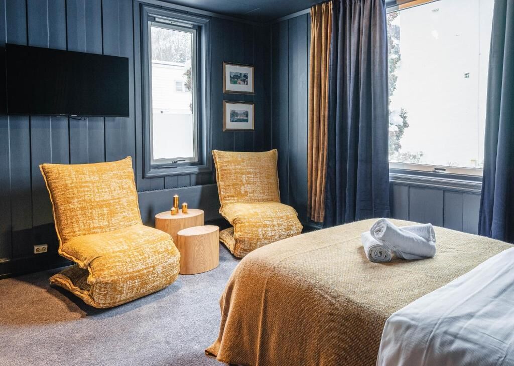 Una habitación de hotel con dos sillones, una cama, un televisor de pantalla plana y un ventanal con cortinas.