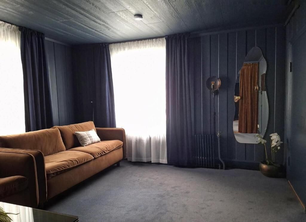 A room with a brown sofa, a window with curtains, a radiator, and a mirror.