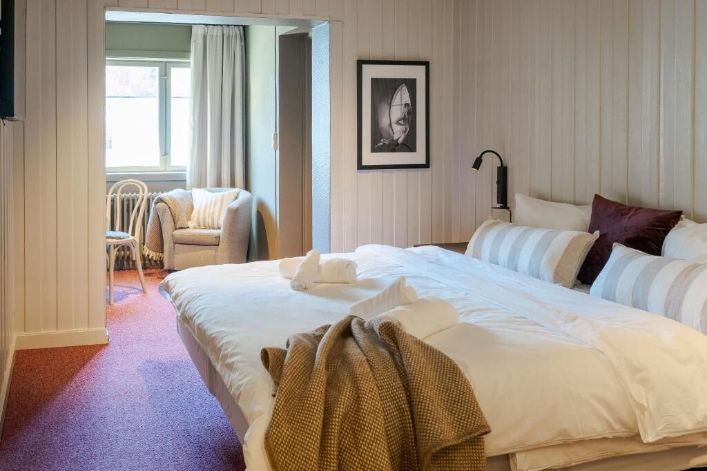 Une chambre d’hôtel avec un grand lit, une chaise, un tableau encadré au mur et une fenêtre avec des rideaux.