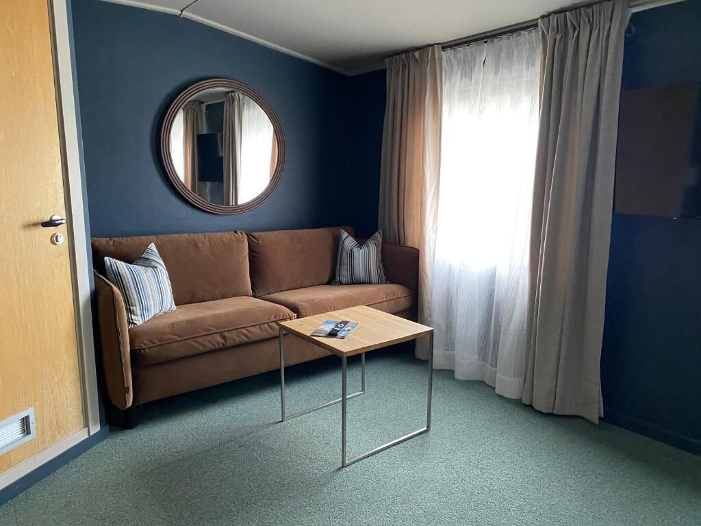 A room with a brown sofa, a round mirror, a small table, and a window with curtains.