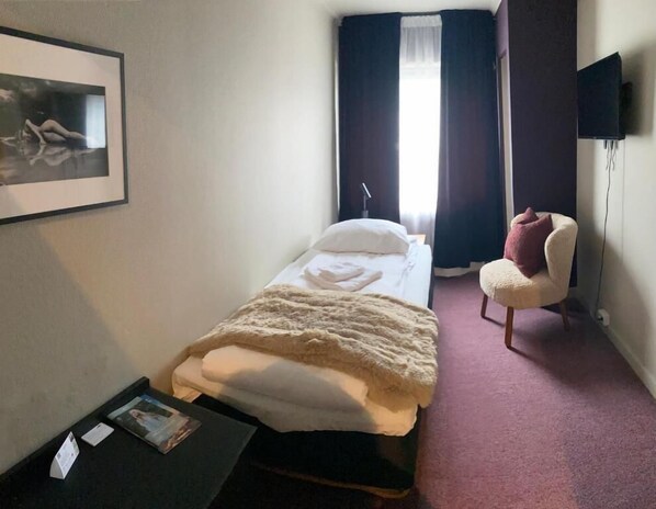 A hotel room with a bed, a chair, a small table, and a wall-mounted TV.