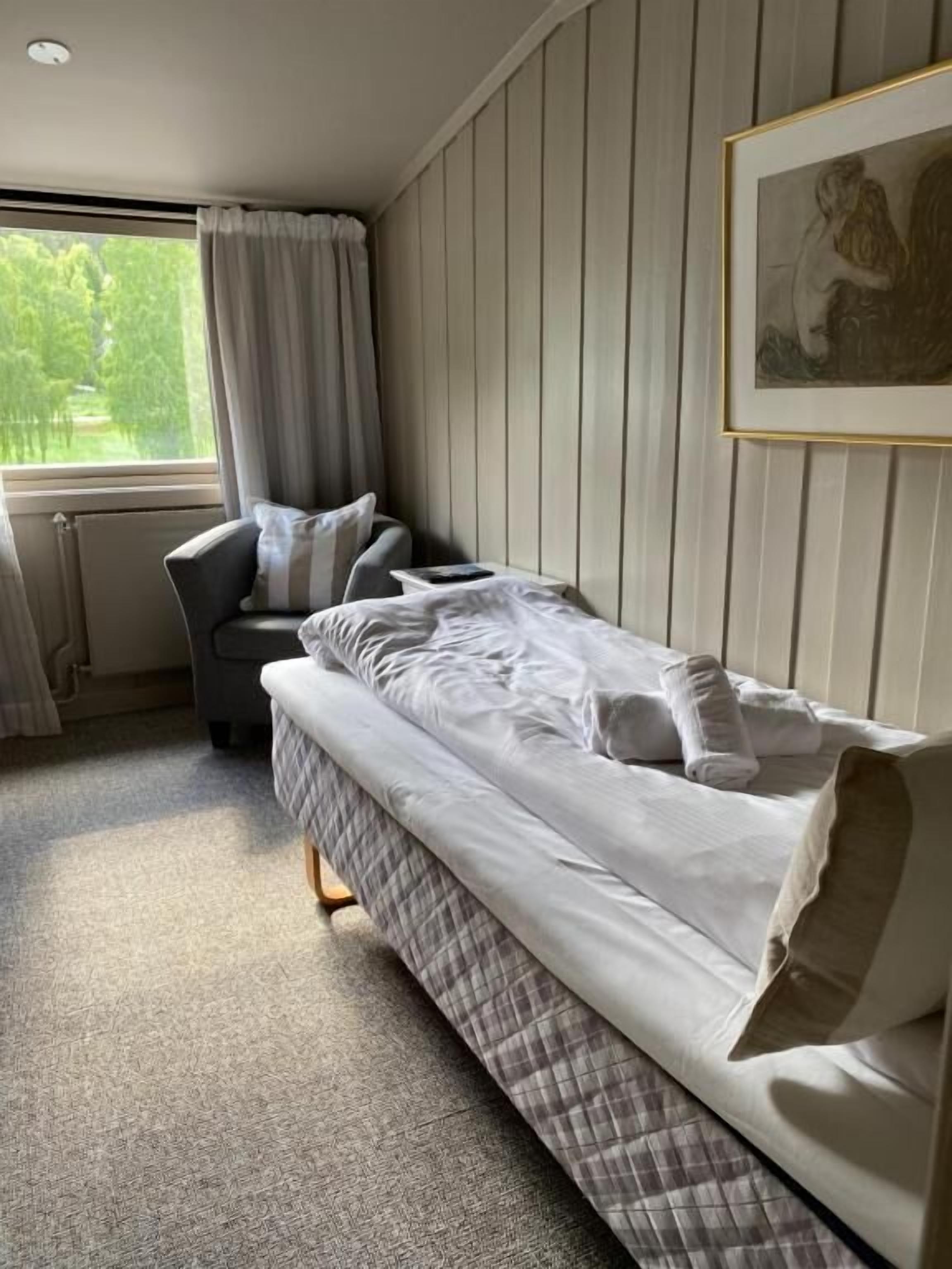 Een enkel bed met hoofdeinde, nachtkastje en stoel in een kamer met raam en een lijst met foto.