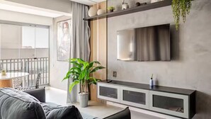 Ein modernes Wohnzimmer mit einer Couch, einem Blumentopf, einem an der Wand montierten Fernseher und einem Balkon mit Tisch und Stühlen.