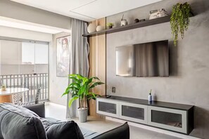 Ein modernes Wohnzimmer mit einer Couch, einem Blumentopf, einem an der Wand montierten Fernseher und einem Balkon mit Tisch und Stühlen.