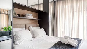 Ein modernes Hotelzimmer mit einem großen Bett, einem Nachttisch, einem Spiegel und Blick durch leichte Vorhänge nach draußen.