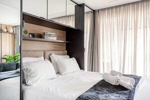 Ein modernes Hotelzimmer mit einem großen Bett, einem Nachttisch, einem Spiegel und Blick durch leichte Vorhänge nach draußen.