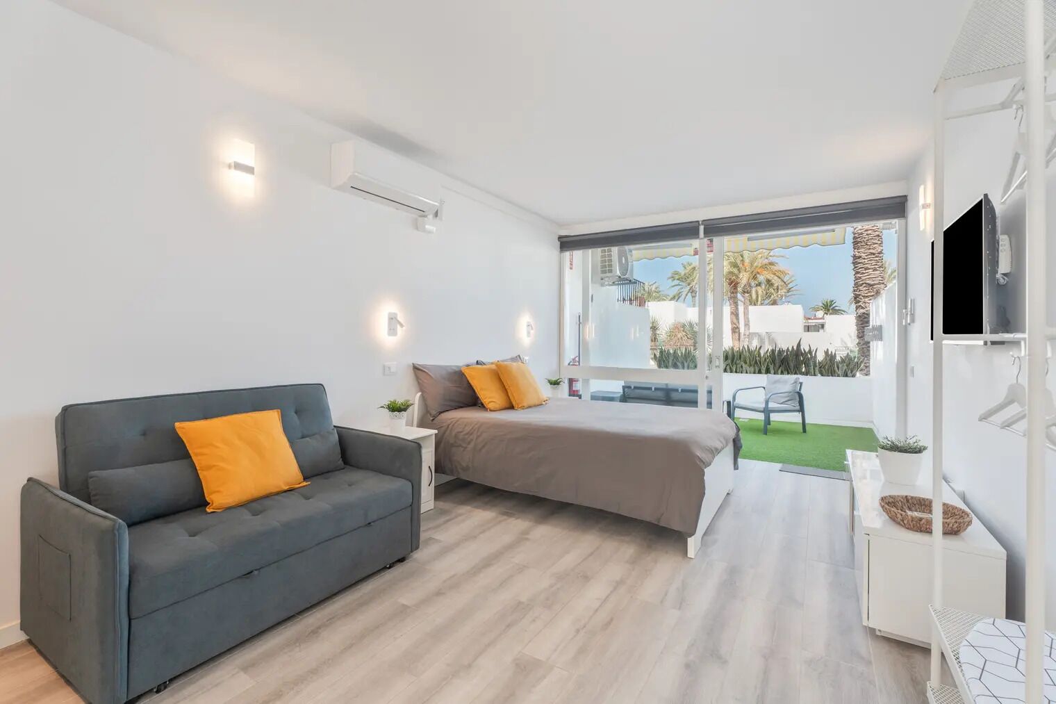 A modern living room with a sofa, a bed, a TV, and a balcony with a view of the outside.