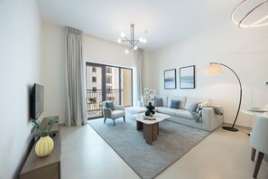 A modern living room with a sofa, armchair, coffee table, and a TV. There is a balcony with a view of buildings.