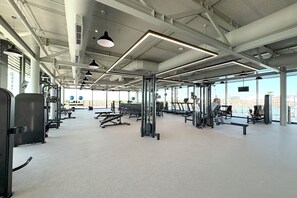 A spacious gym with various exercise equipment, large windows, and a high ceiling.