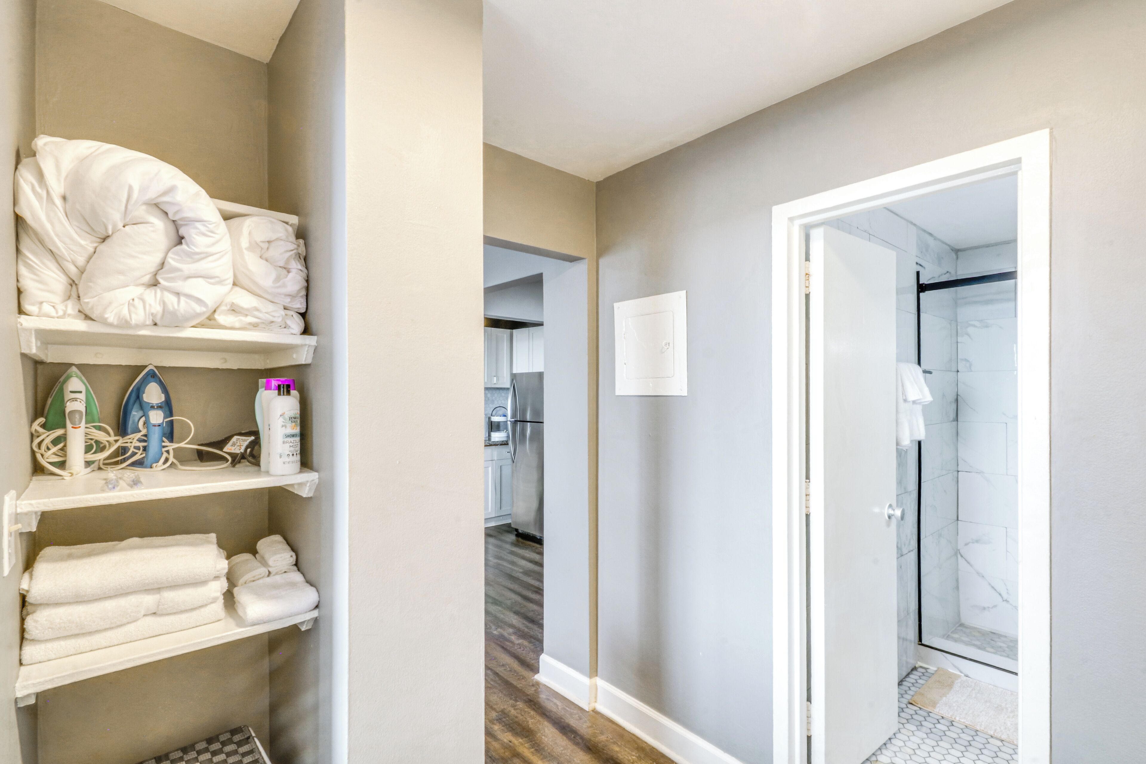 A hotel room with a shower, a hallway leading to a kitchen, and a storage area with towels and toiletries.