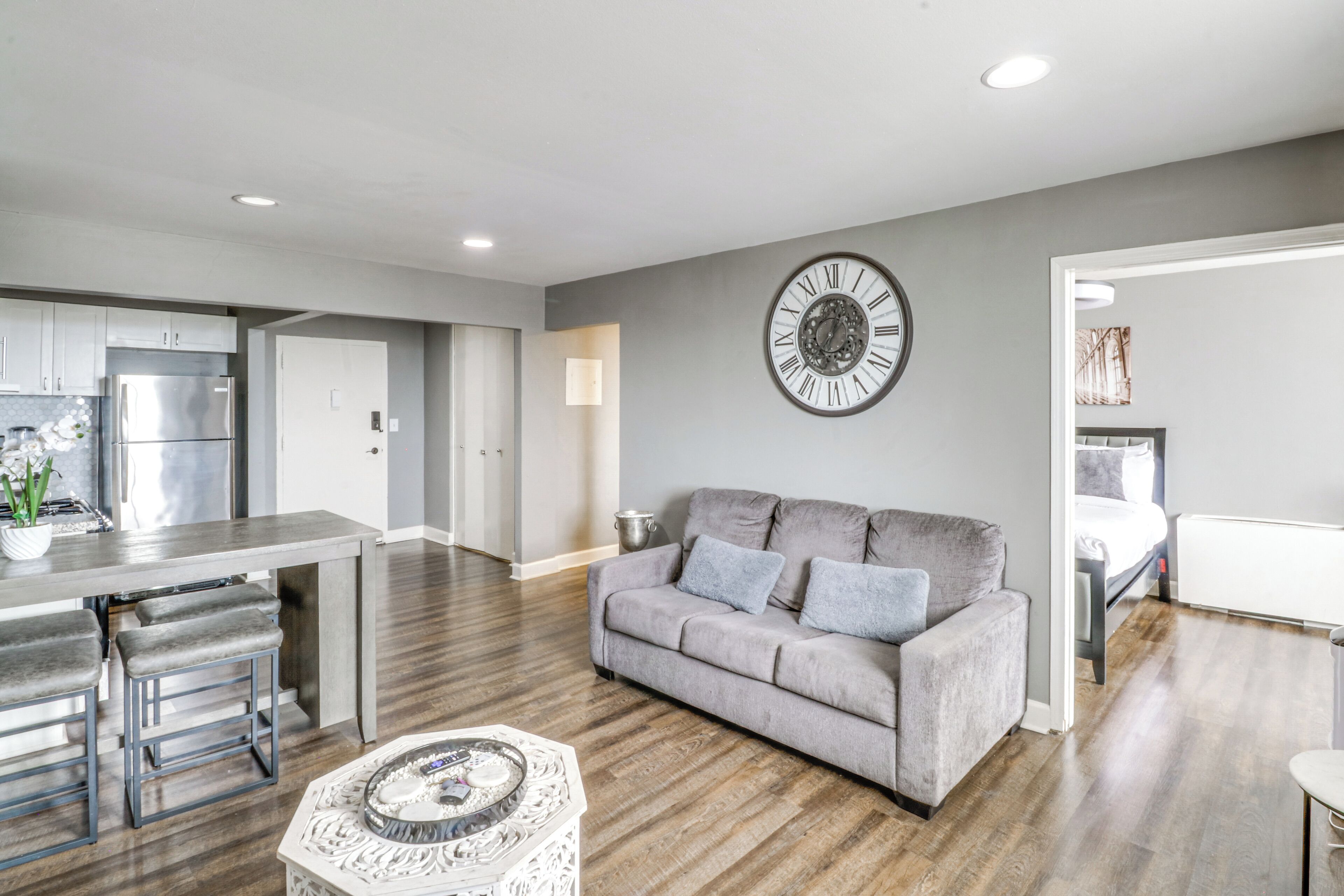 A modern living room with a grey sofa, a decorative clock, and a dining area with a kitchen in the background.