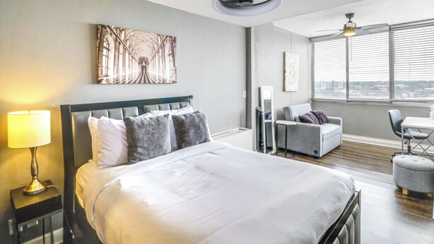 A hotel room with a bed, a desk, a chair, a lamp, and a large window with blinds. - Downtown Atlanta High-rise Condo (Atlanta)