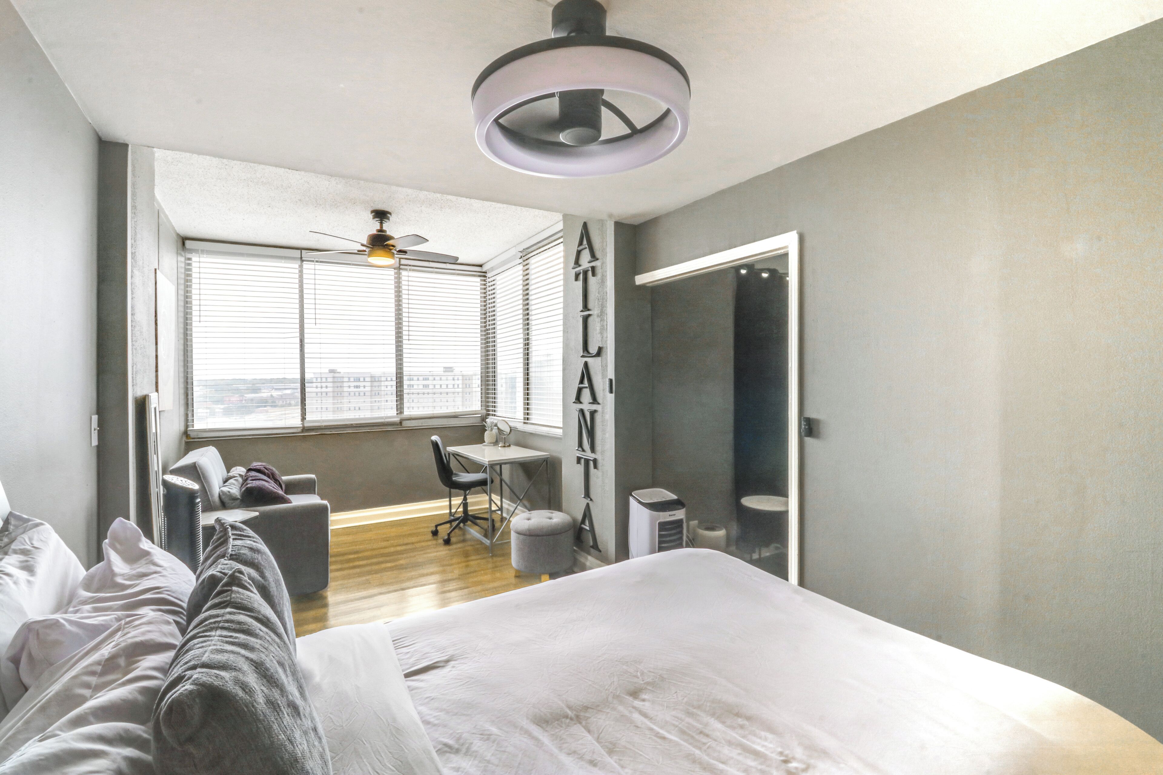 A modern bedroom with a large bed, a desk, and a chair. There is a window with blinds, a ceiling fan, and a view of the city.