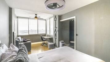 A modern bedroom with a large bed, a desk, and a chair. There is a window with blinds, a ceiling fan, and a view of the city.