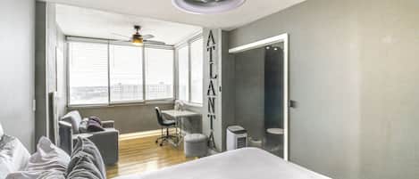 A modern bedroom with a large bed, a desk, and a chair. There is a window with blinds, a ceiling fan, and a view of the city.