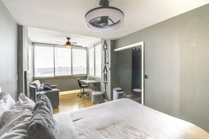 A modern bedroom with a large bed, a desk, and a chair. There is a window with blinds, a ceiling fan, and a view of the city. - Downtown Atlanta High-rise Condo (Atlanta)