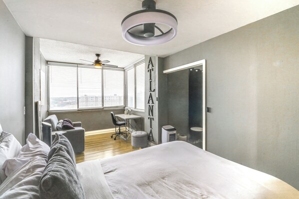 Un dormitorio moderno con una cama grande, un escritorio y una silla. Hay una ventana con persianas, un ventilador de techo y vistas a la ciudad.