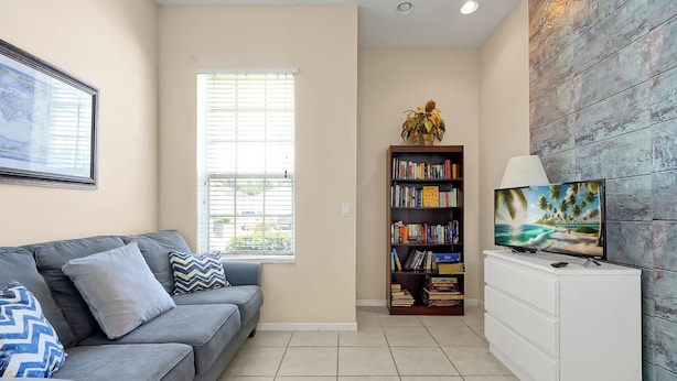 A living room with a grey sofa, a bookshelf, a TV, and a window with blinds. - 4BR Golf Villa w/ Pool, Near Disney & Beaches (Davenport)