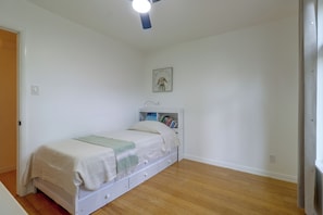 Ein Schlafzimmer mit einem Einzelbett, einem Deckenventilator, einem gerahmten Bild an der Wand und einem Bücherregal mit Büchern.