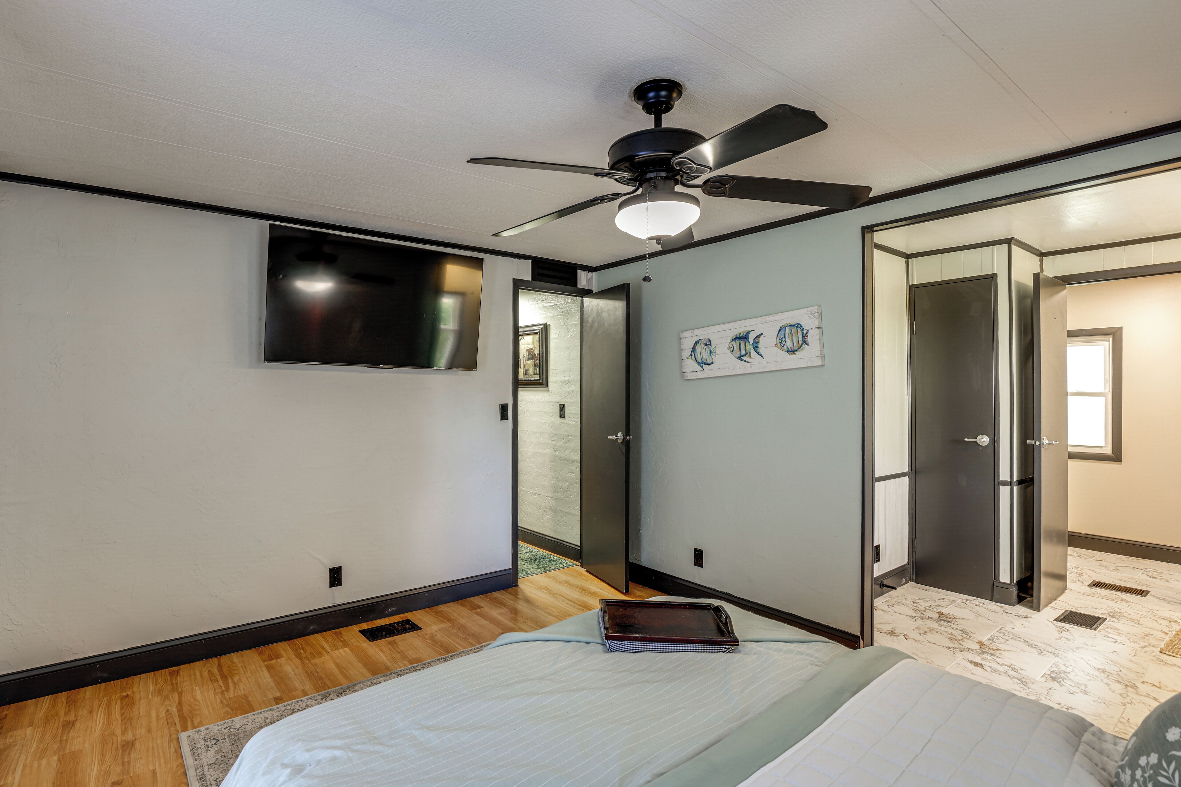 A bedroom with a bed, a ceiling fan, a flat-screen TV, a door, and a wall with a fish painting.