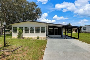 A single-story building with a covered porch, a front door, and a small front yard with grass and a tree. - 4-passenger Golf Cart! Fun in the Villages (Lady Lake)