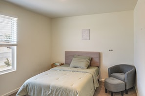 A bedroom with a bed, a chair, a bedside table, and a window with blinds.