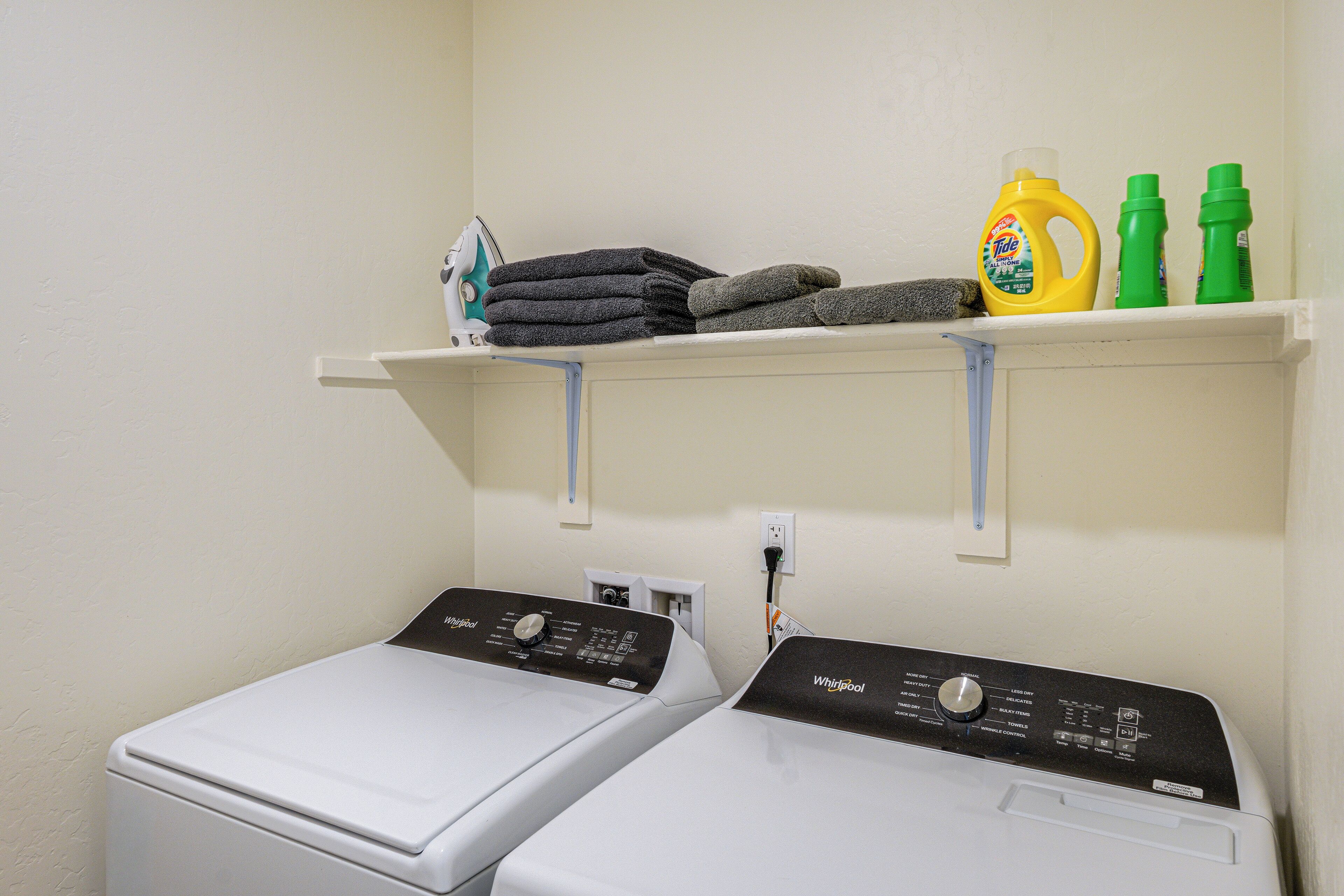 A laundry area with two front-loading washers, a shelf with folded towels and laundry supplies, and a power outlet.