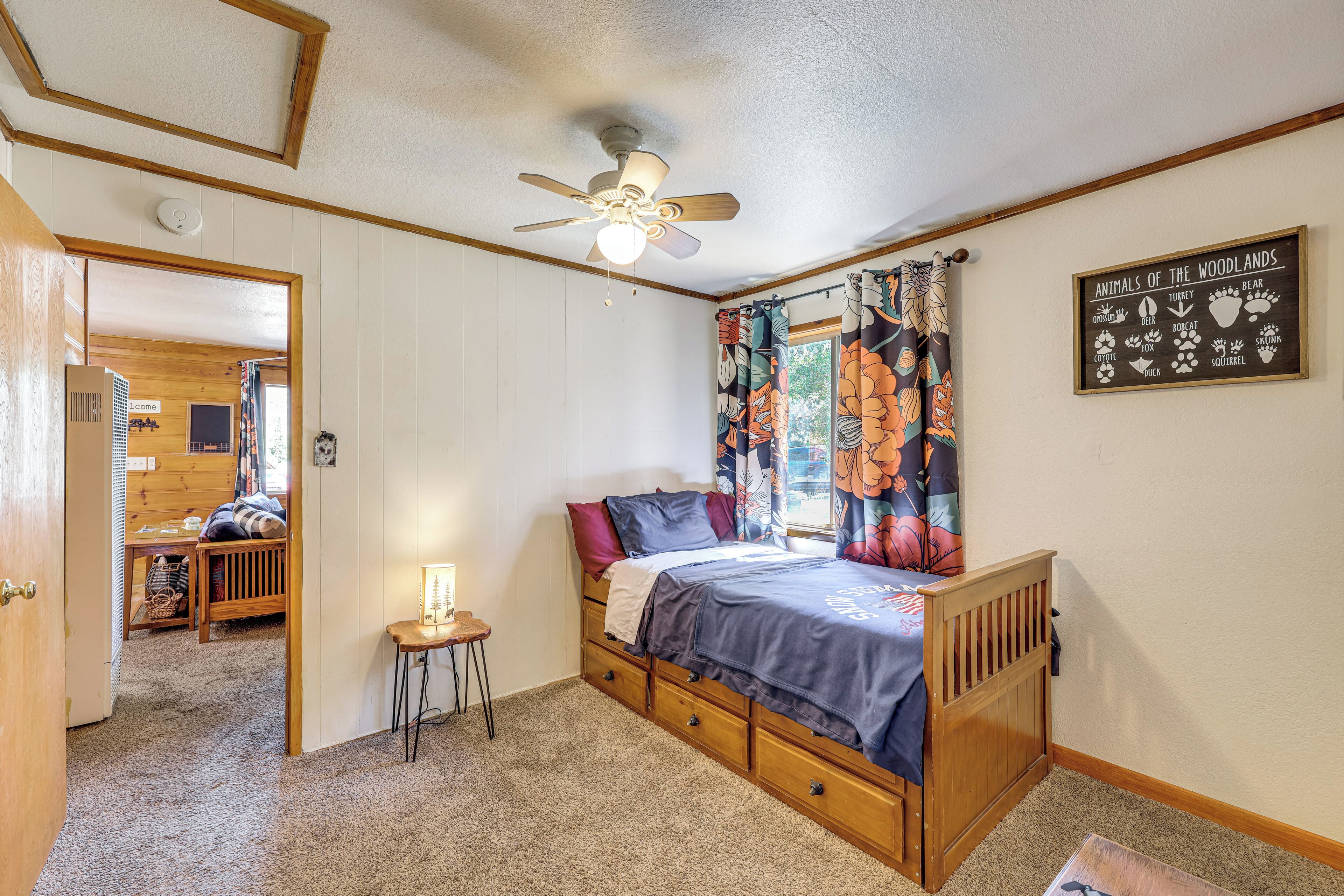 A bedroom with a wooden bed, a ceiling fan, a small table with a lamp, and a window with floral curtains.