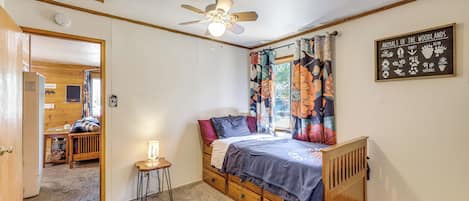 Un dormitorio con cama de madera, ventilador de techo, una mesita con lámpara y ventana con cortinas florales.